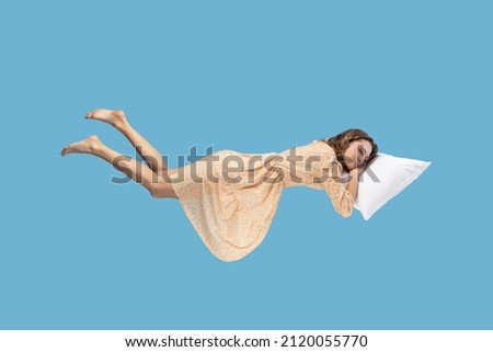 Relaxed girl in yellow dress levitating in mid-air, sleeping on stomach lying comfortable cozy on pillow, keeping eyes closed, watching peaceful dream. indoor studio shot isolated on blue