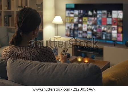 Woman relaxing on the couch, she is using the remote control and choosing a TV show or movie on the television menu Royalty-Free Stock Photo #2120056469