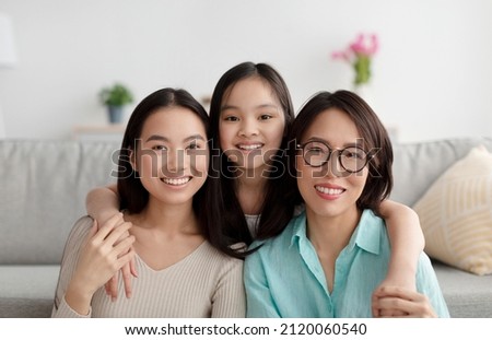 Portrait of joyful Asian family of mature mother, adult daughter and little granddaughter smiling at camera at home. Millennial woman with her child and mom having friendly relations indoors Royalty-Free Stock Photo #2120060540