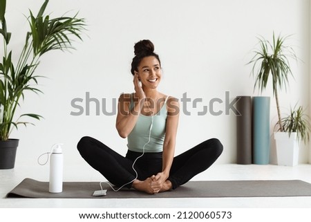 Beautiful Young Athletic Woman In Earphones Listening Music On Smartphone While Exercising In Yoga Studio, Cheerful Sporty Female Sitting On Fitness Mat, Enjoying Favorite Playlist For Trainings