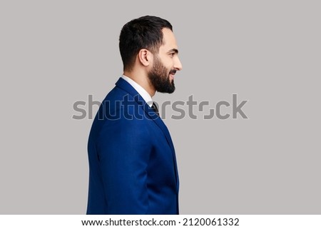 Side view portrait of cheerful bearded man with smile, standing and looking at camera, expressing positive emotions, wearing official style suit. Indoor studio shot isolated on gray background. Royalty-Free Stock Photo #2120061332