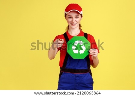 Portrait of satisfied worker woman standing and holding recycling sign, thinking green, pointing at camera, wearing overalls and red cap. Indoor studio shot isolated on yellow background.