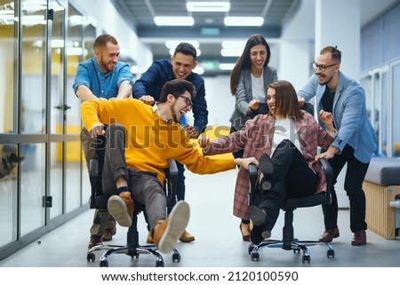 Young cheerful business people  having fun while racing on office chairs and smiling. Friendly work team enjoying fun work break activities.