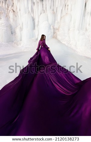 Art fantasy beautiful woman posing in a purple purple flying dress, with a long train of fabric. Behind her is a winter frozen shore covered in blue and white ice. The model stands with her back Royalty-Free Stock Photo #2120138273