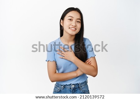 Smiling humble asian girl, hold hands on chest and looking grateful, appreciate smth, standing over white background Royalty-Free Stock Photo #2120139572
