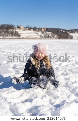 Young caucasian smiling preschool girl sitting in warm clothes throws snow up playing and having fun on sunny winter weather outside on snowy field in park. Christmas holidays, childhood, carefree