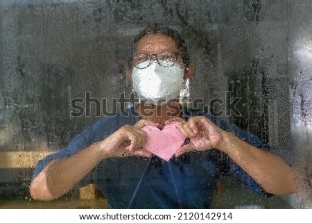 A woman wearing mask who is sitting behind a glass window gesturing love with her hands and a heart-shaped pink paper.