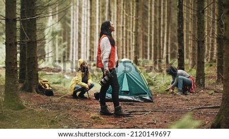 Young woman in outdoor gear taking photos with a telephoto lens while camping with friends in a forest