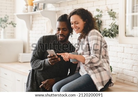 Middle-aged mature happy african couple using smart phone cellphone for surfing social media, sharing photos, checking e-mail, using mobile application online relaxing at home kitchen Royalty-Free Stock Photo #2120200091