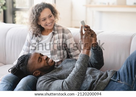 Middle-aged mature happy african couple using smart phone cellphone for surfing social media, sharing photos, checking e-mail, using mobile application online relaxing at home on sofa