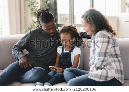 Little preteen african-american girl daughter using smart phone cellphone, surfing social media online with her parents relaxing on the couch in living-room at home. Mobile application
