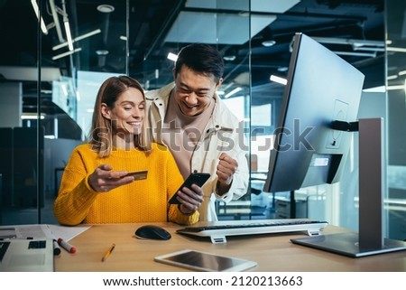 Creative team of Asian man and woman shopping online, sitting at a table, choosing a gift together, rejoicing and smiling, using a smartphone and credit card