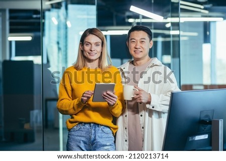 a team of two Asian male and female workers take a break, look at the camera and smile, use a tablet to discuss a joint business project