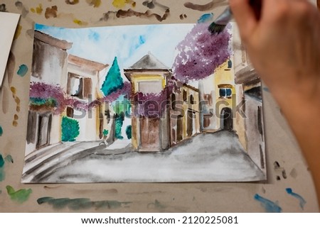 Closeup of vibrant watercolor painting process. Brush in hand above painting. Colorful medieval city street on paper board.
