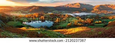 Wide panoramic view of small lake and mountains with beautiful golden sunrise light on landscape. Loughrigg Tarn, Lake District, UK.