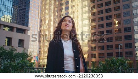 Cute girl walking alone quiet city street on holiday. Happy brunette woman looking around on cityscape summer day. Young asian lady enjoy modern buildings view. Active leisure in big town weekend. Royalty-Free Stock Photo #2120238053