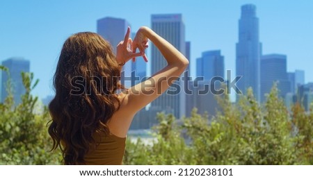 Unknown girl fingers make sign LA on blurred cityscape view. Unrecognizable brunette standing in green park showing name town Los Angeles by hands closeup. Stylish woman tourist relax on nature.