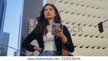 Beautiful brunette girl using smartphone standing on street close up. Young confident woman looking screen white smart phone outdoors. Attractive asian lady typing on modern phone in front skyscrapers Royalty-Free Stock Photo #2120238296