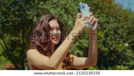 Cheerful asian girl photographing beautiful landscape in sunny park closeup. Happy smiling woman making photo using modern smartphone outdoors. Attractive brunette taking picture summer nature.