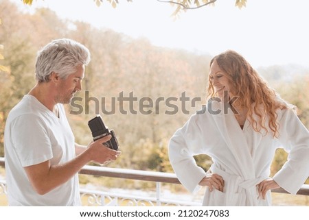 Man with old-fashioned camera photographing woman in bathrobe on autumn balcony