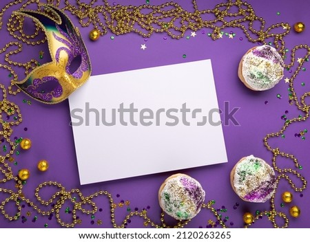 Mardi Gras King Cake sufganiyot donuts, masquerade festival carnival mask, gold beads and golden, green, purple confetti on purple background. Holiday party invitation, greeting card concept. Top view