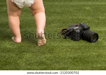 A modern camera lies on the grass next to the bare feet of a small child. The concept of a young photographer, parenthood, childhood.