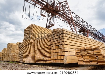 Stacked lumber in the warehouse of the production site against the background of a cantilever gantry crane. Royalty-Free Stock Photo #2120403584