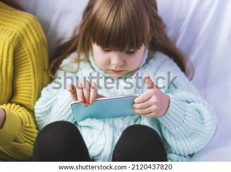 A beautiful little girl is watching cartoons on her smartphone online. A small baby girl is playing a game on a mobile phone. I am interested in modern technologies. Gadgets and kids. Leisure at home.