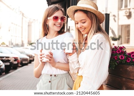 Two young beautiful smiling hipster female in trendy summer clothes.Sexy carefree women posing in the street. Positive pure models having fun at sunset. They drinking coffee or tea in plastic cup Royalty-Free Stock Photo #2120445605
