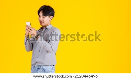 Studio shot of millennial Asian thoughtful doubtful curious male fashion model in stylish fashionable casual outfit standing holding  smartphone on yellow background.