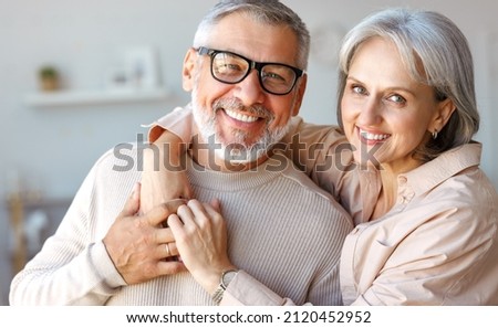 Close up of beautiful smiling senior family couple husband and wife looking at camera with tenderness and love while standing in living room at home, retired man and woman embracing indoors Royalty-Free Stock Photo #2120452952