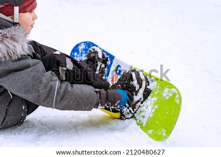 little boy sitting on snow putting his feet in snowboard bindings adjusting straps. High quality photo Royalty-Free Stock Photo #2120480627