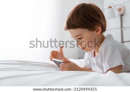 Little baby boy lays on his bed and plays with a smartphone. Close-up portrait. Holds in hands black cell phone. Concept of online education. White room and clothes. Telephone user. Save child vision