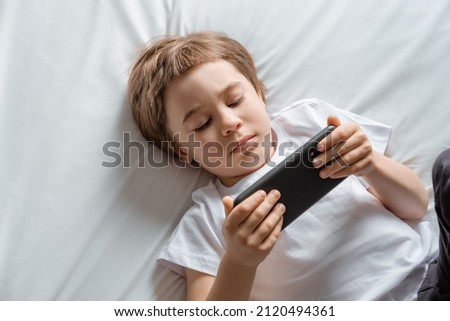 Little baby boy lays on his bed and plays with a smartphone. Close-up portrait. Holds in hands black cell phone. Concept of online education. White room and clothes. Telephone user. Save child vision