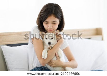 Portrait closeup shot of Asian young happy pretty teenager girl in casual outfit sitting leaning at pillow smiling look at camera on bed holding hugging white small Shiba Inu puppy dog in bedroom.