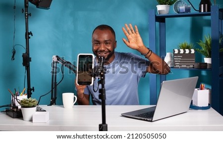 Content creator doing greeting hand gesture looking at live video podcast setup sitting at desk with professional microphone. Smiling vlogger waving hello in front of recording smartphone.