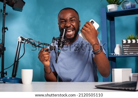 Influencer screaming after reading on smartphone insulting comments from online social media account. Portrait of vlogger sitting at desk in vlogging studio deeply hurt by online bully.