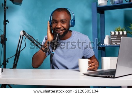 African american vlogger pressing wireless headphones to ear with hand listening to fan talking while holding white cup. Influencer sitting at desk with laptop interacting with audience.