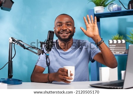 Content creator waving hand to followers while holding white cup in online morning show in front of laptop computer. Vlogger saying hello at audience sitting at desk with professional microphone.