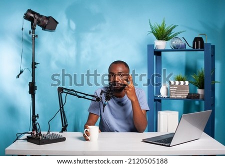 Vlogger showing peace sign sitting while holding cup at desk in recording studio with professional microphone and video light. Content creator doing hand gesture in front of audio podcast setup.