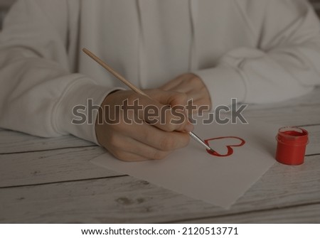 A girl draws a red heart with gouache on a white sheet of paper. The girl draws with red gouache on paper. Royalty-Free Stock Photo #2120513771