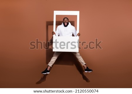 Photo of excited funky guy wear white hoodie jumping high holding paper frame isolated brown color background