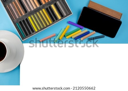 Flat lay soft dry artist pastels, white paper sheet,black smartphone by side,coffee cup on saucer.Online drawing with pastel crayons on blue.Relaxation stress relief through hobby concept.Copy space.