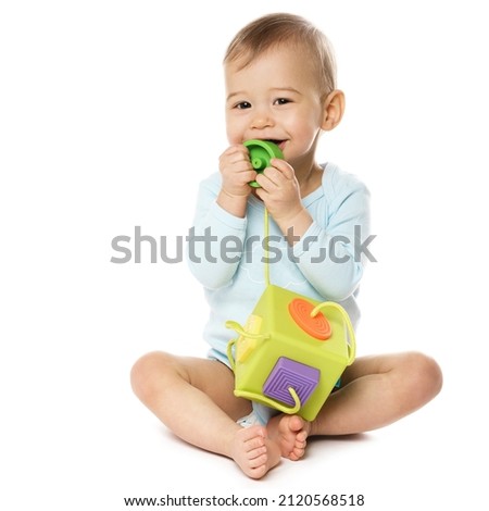 Adorable smiling little boy in a romper is sitting and playing with plastic toy, putting it in his mouth on white background. Royalty-Free Stock Photo #2120568518