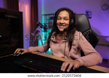 Portrait of a smiling young gamer, the girl is happy to win the game, pass the level, the teenager has a headset, professional gaming accessories, the room lit by fioket blue led lights