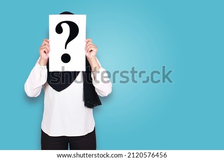 Who am I concept. Anonymouse muslim lady wearing hijab covering her face with paper of question mark, against blue background Royalty-Free Stock Photo #2120576456