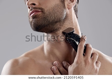 Obscure face of young man shaving beard with electric razor. Handsome dark-haired bearded guy. Concept of face care. Idea of male beauty. Isolated on white background. Studio shoot Royalty-Free Stock Photo #2120591405