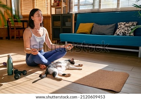 Woman practicing yoga and meditation at home sitting on yoga mat in lotus pose with her cat, stretching muscles of legs. Mindful meditation concept. Wellbeing. Royalty-Free Stock Photo #2120609453