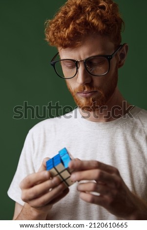 Young focused caucasian clever red hair guy playing rubik's cube in hands. Concept of human of generation Z. Handsome bearded male person in glasses. Isolated on dark green background in studio Royalty-Free Stock Photo #2120610665