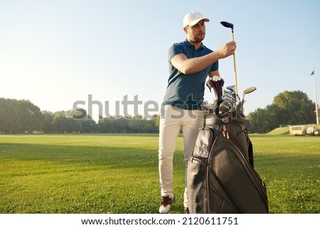 Golfer choosing putter before playing golf on green lawn at sunny day. Concept of entertainment, recreation, leisure and hobby outdoors. Young caucasian man wearing cap, t-shirt, pants and shoes Royalty-Free Stock Photo #2120611751